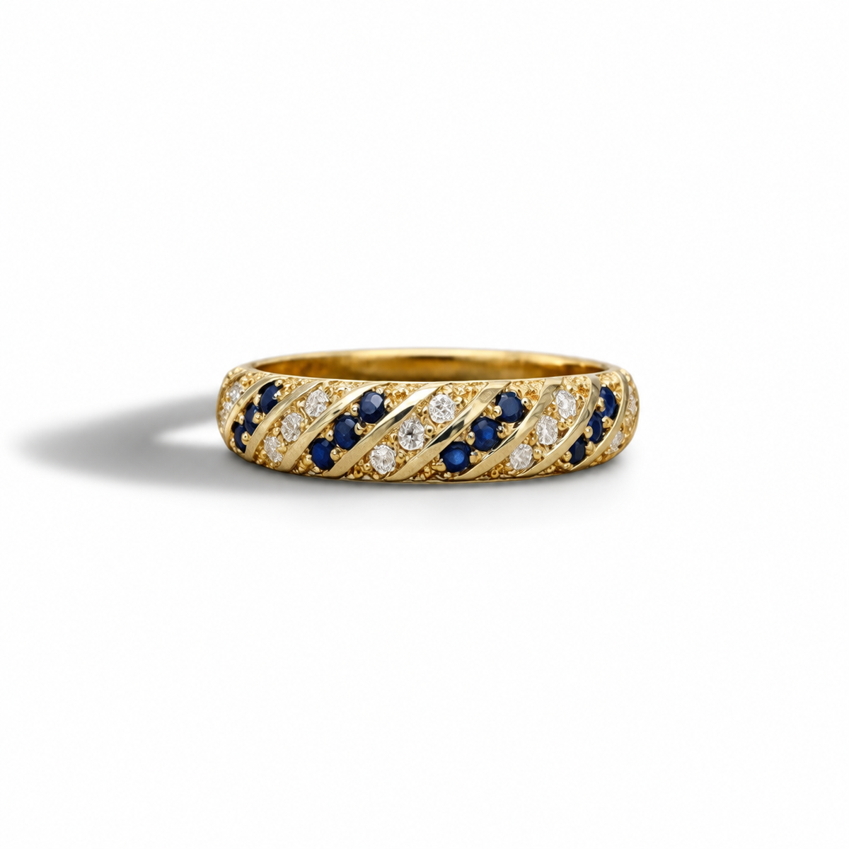 A yellow gold eternity ring featuring diagonal bands of round-cut dark blue sapphires and clear pavé diamonds, set against a clean white background with a soft shadow.