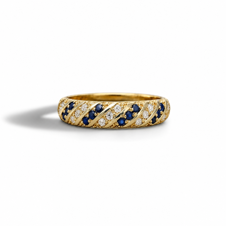 A yellow gold eternity ring featuring diagonal bands of round-cut dark blue sapphires and clear pavé diamonds, set against a clean white background with a soft shadow.