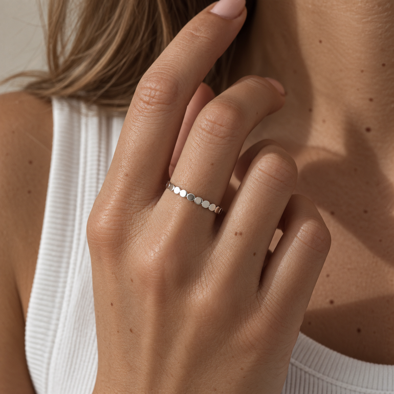 A close-up of a woman's hand wearing a delicate sterling silver band ring with small, evenly spaced round-cut stones, set against a soft beige background.