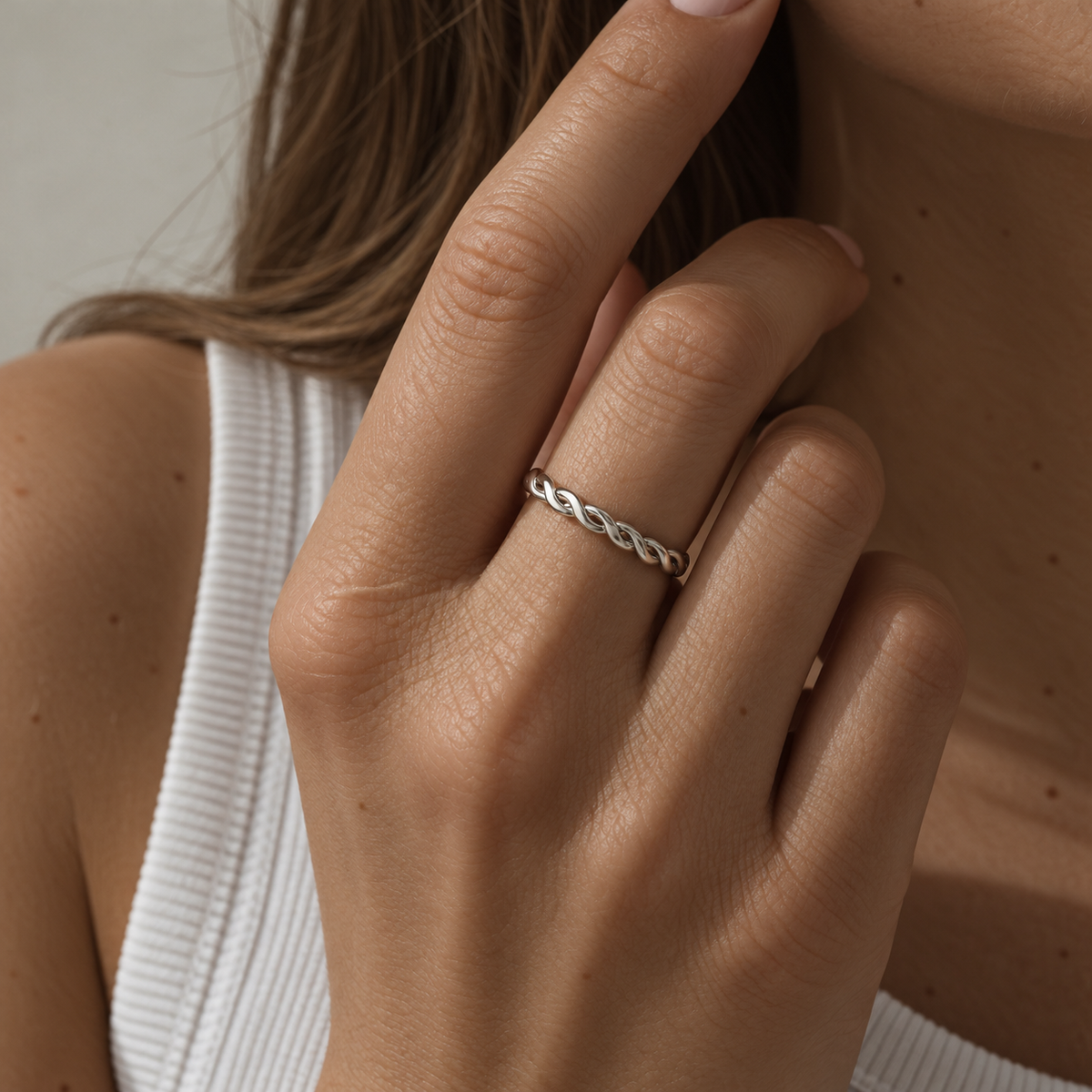 A close-up of a woman's hand wearing a thin silver band ring with an intricate twisted or braided design, paired with a white ribbed tank top.