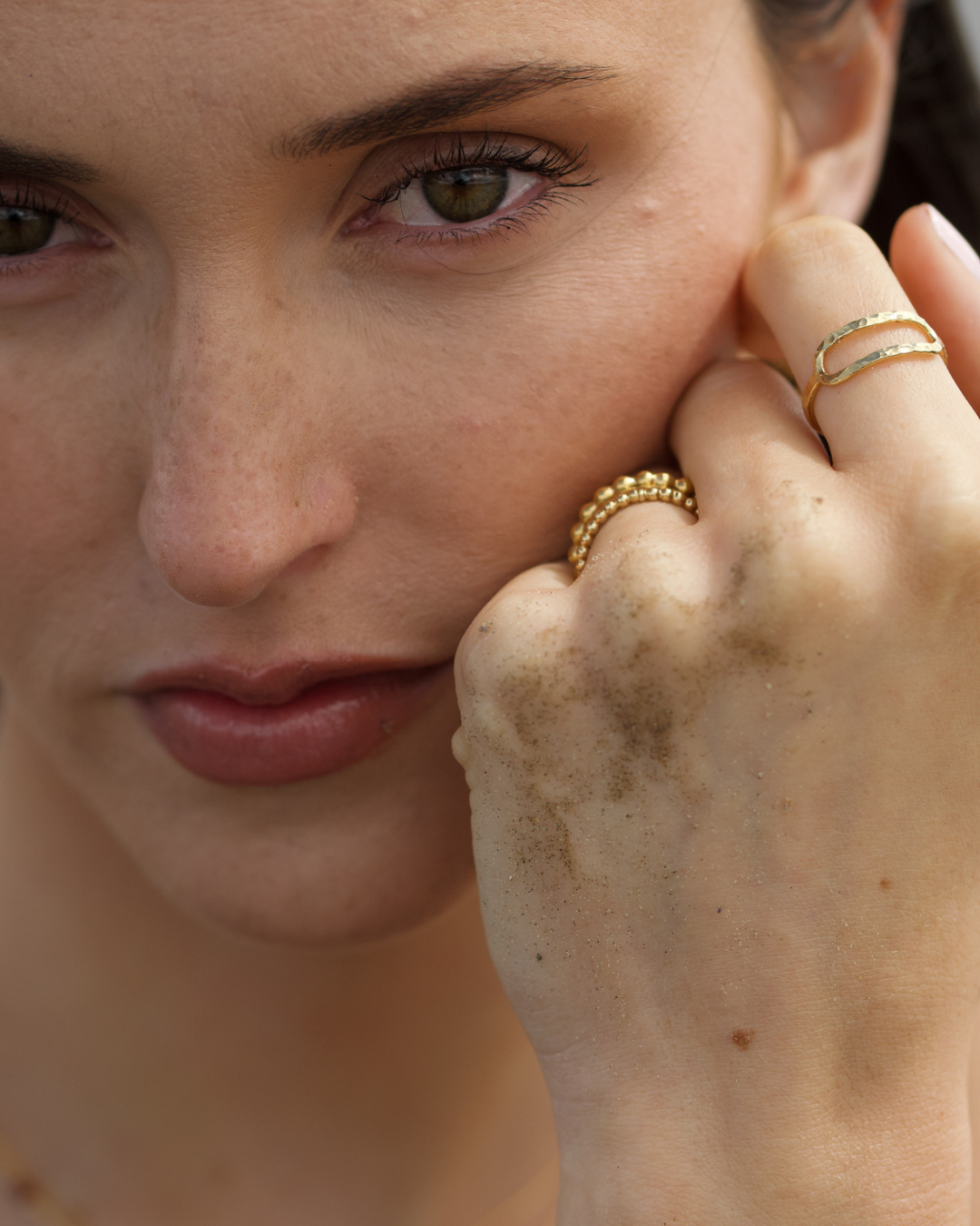 Close-up of a woman resting her face on her hand, showcasing three gold rings: a textured beaded ring, a thin double-band ring, and a delicate gold chain ring.