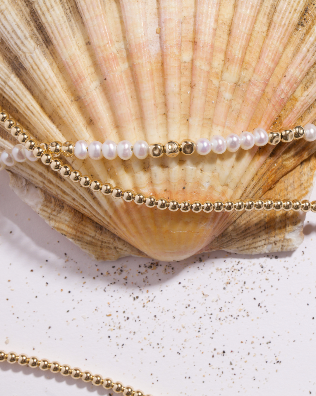 Gold beaded chains and a strand of white pearls draped over a large beige scallop shell on a white surface with scattered sea salt.