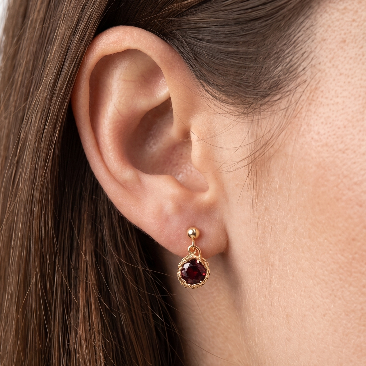 Close-up of a person wearing a gold earring with a red gemstone on a neutral background