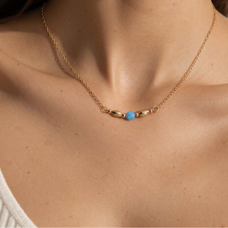 A close-up of a model's neck and upper chest wearing a delicate gold chain necklace with a small central blue bead accent.