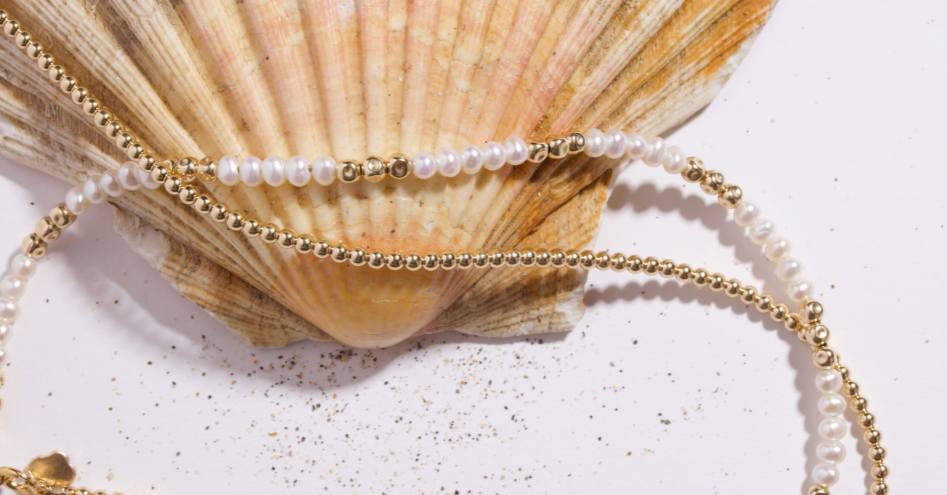 A close-up of a double-strand necklace featuring white faux pearls and polished gold beads, draped over a large beige seashell on a white surface.