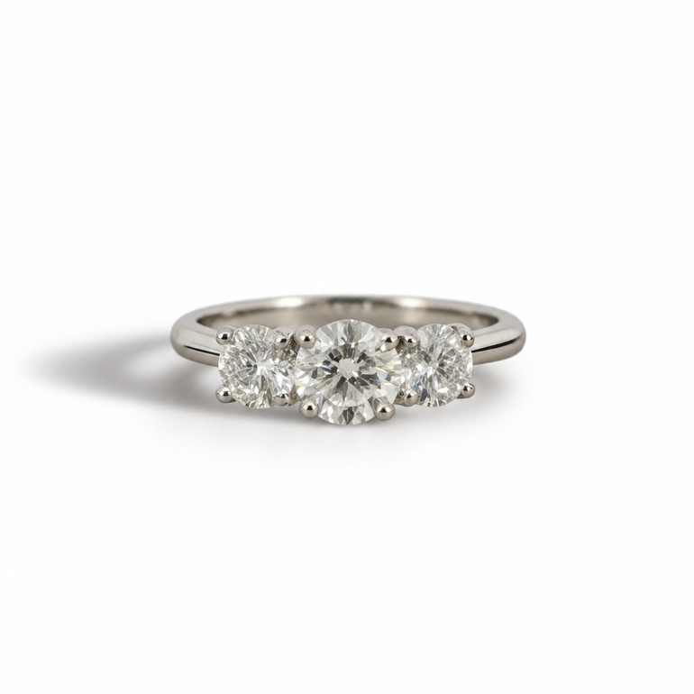 A three-stone diamond ring in a silver-colored metal band platinum , featuring a round center diamond flanked by two round side diamonds, set against a plain white background.